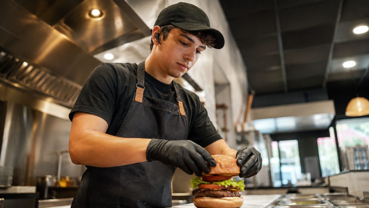 A QSR kitchen worker wearing an earpiece while assembling a burger at a prep station